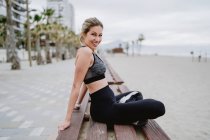 Side view of concentrated lady athlete sitting on bench and looking in camera with seaside on blurred background — Stock Photo