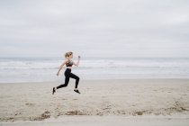 Vue latérale du joggeur féminin déterminé en vêtements de sport et baskets élégants sautant au bord de la mer vide par temps nuageux — Photo de stock