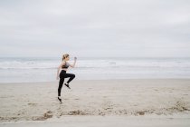 Vista laterale di determinata jogger femminile in elegante abbigliamento sportivo e scarpe da ginnastica che saltano a riva vuota sul tempo nuvoloso — Foto stock