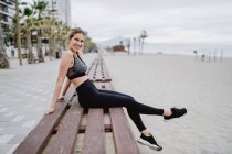 Side view of concentrated athlete in trendy active wear sitting on bench and looking in camera on seaside — Stock Photo