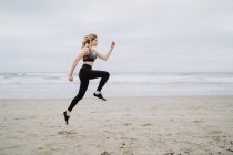 Side view of determined female jogger in stylish sportswear and sneakers jumping at empty seashore on cloudy weather — Stock Photo