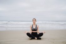 Молодая стройная женщина в черном топе и леггинсы, сидящие в позе лотоса на пляже с закрытыми глазами во время медитации — стоковое фото