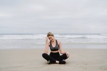 Giovane donna magra in top nero e leggings seduti in posizione di loto e sorridente in spiaggia — Foto stock