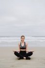 Giovane donna magra in top nero e leggings seduti in posizione loto mentre medita in spiaggia — Foto stock