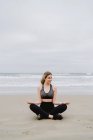 Jeune femme mince en haut noir et leggings assis en position lotus et regardant loin de la plage — Photo de stock