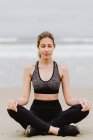 Giovane donna magra in top nero e leggings seduti in posizione loto in spiaggia con gli occhi chiusi mentre medita — Foto stock