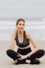 Junge schlanke Frau in schwarzem Top und Leggings sitzt in Lotusposition, während sie am Strand meditiert — Stockfoto