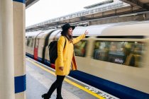 Side view of happy stylish female smiling and waiving hand near train while standing on platform on station in London, United Kingdom — Stock Photo