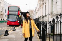 Молода жінка в модному жовтому пальто захоплюється декоративними будівлями, стоячи на тротуарі біля подвійного декера на вулиці Лондона, Велика Британія. — стокове фото