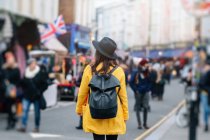 Back view of woman in yellow coat and stylish hat walking on road during carnival in London, United Kingdom — Stock Photo