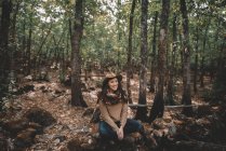 Calme jeune femme heureuse dans un chapeau élégant et écharpe assis sur le sol regardant loin tout en se reposant dans la forêt verte — Photo de stock