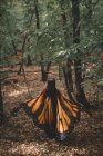 Анонимная молодая женщина в плаще с крыльями бабочки танцует возле деревьев в зеленом лесу — стоковое фото