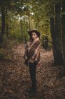 Боковой вид молодой женщины в шляпе, завернутой в клетчатый шарф, стоя на сухих листьях осеннего леса — стоковое фото
