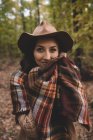 Jeune femme en chapeau enveloppant dans une écharpe à carreaux tout en se tenant debout sur des feuilles sèches dans la forêt d'automne regardant à la caméra — Photo de stock