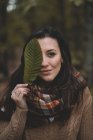 Stylish lady in knitted sweater and checkered scarf covering eye with leaf while standing on blurred background of forest — Stock Photo