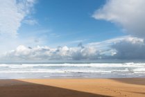 Idílica playa de arena vacía a lo largo del océano turquesa con olas y espuma bajo el cielo azul nublado en Zarautz en España - foto de stock