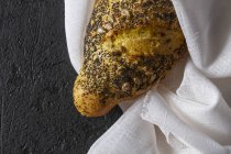 Loaf of ruddy crispy fresh bread with grains and poppy seeds wrapped in towel on grey background — Stock Photo