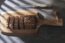 Sliced loaf of wholegrain bread on wooden cutting board and kitchen knife placed on wooden table — Stock Photo