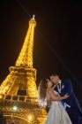 Freudiger Bräutigam im blauen Anzug und Braut im weißen Brautkleid umarmen sich lächelnd und küssen sich am Abend mit Eiffelturm im Hintergrund in Paris — Stockfoto