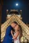 Молодая супружеская пара в свадебном костюме и платье обнимаются, стоя на качающейся арке с Лувром на заднем плане в Париже — стоковое фото