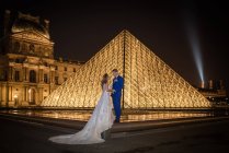 Young newly married couple in wedding suit and gown hugging while standing in rocked arch with Louvre on background at Paris — Stock Photo