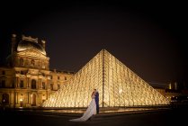 Young newly married couple in wedding suit and gown hugging while standing in rocked arch with Louvre on background at Paris — Stock Photo