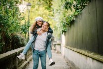 Content woman in casual clothing sitting on back and kissing pleased man walking on small alley with green plants on blurred background — Stock Photo