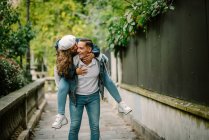 Content woman in casual clothing sitting on back and kissing pleased man walking on small alley with green plants on blurred background — Stock Photo