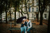 Mujer contenta con ropa casual sentada con un joven en el banco de un hermoso vecindario sosteniendo un paraguas - foto de stock