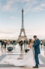 Novio en traje azul y novia en vestido de novia blanco teniendo baile lento sonriendo y mirándose el uno al otro con la Torre Eiffel en el fondo en París - foto de stock