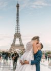 Groom in blue suit and bride in white wedding gown kissing passionately with Eiffel Tower on background at Paris — Stock Photo
