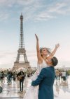 Bräutigam im blauen Anzug hebt fröhliche Braut mit offenen Armen im weißen Hochzeitskleid mit Eiffelturm auf Hintergrund in Paris — Stockfoto