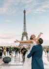 Bräutigam im blauen Anzug hebt fröhliche Braut mit offenen Armen im weißen Hochzeitskleid mit Eiffelturm auf Hintergrund in Paris — Stockfoto