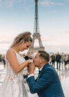 Novio contenido en traje elegante azul de pie sobre la rodilla y besar las manos de la novia satisfecha en vestido de novia blanco con Torre Eiffel en el fondo - foto de stock