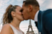 З фокусу наречений в синьому костюмі і наречена в білій весільній сукні цілуються пристрасно з Ейфель Тауер на задньому плані в Парижі — стокове фото
