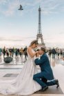 Zufriedener Bräutigam in blauem stylischem Anzug auf Knien stehend und küssende Hände einer zufriedenen Braut im weißen Hochzeitskleid mit Eiffelturm im Hintergrund — Stockfoto