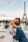 Zufriedener Bräutigam in blauem stylischem Anzug steht auf Knien und küsst zufriedene Braut im weißen Hochzeitskleid mit Eiffelturm im Hintergrund — Stockfoto