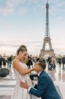 Novio en traje azul arrodillado y novia en vestido de novia blanco abrazándose con la Torre Eiffel en el fondo en París - foto de stock