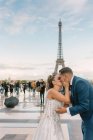 Groom en costume bleu agenouillé et mariée en robe de mariée blanche embrassant passionnément avec Tour Eiffel sur fond à Paris — Photo de stock