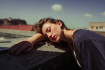 Modelo feminino jovem e sem emoções com lábios vermelhos em roupas casuais apoiadas em cerca balançada com céu azul no fundo desfocado no telhado dos edifícios — Fotografia de Stock