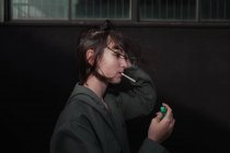 Вид сбоку на юную брюнетку-бунтаря в стильной куртке с синими ногтями, закуривающую сигарету на городской улице — стоковое фото