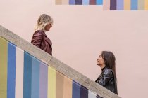 Side view of attractive pensive young friends in jackets standing on different level of striped colored stairs outdoors looking at each other — Stock Photo