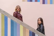 Side view of attractive pensive young friends in jackets standing on different level of striped colored stairs outdoors looking at camera — Stock Photo
