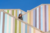 Vue latérale de la jeune femme attrayante et coûteuse en vestes debout sur différents niveaux d'escaliers colorés rayés à l'extérieur regardant loin — Photo de stock