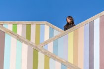 Вид на привлекательную задумчивую молодую женщину в куртках, стоящую на разных уровнях полосатой цветной лестницы на открытом воздухе и смотрящую в сторону — стоковое фото