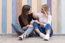 Smiling positive elegant women leaning on each other while sitting close on sidewalk near striped street wall looking at each other — Stock Photo