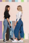 Smiling positive elegant women standing while leaning on stripped colorful wall close to sidewalk on the street looking at each other — Stock Photo