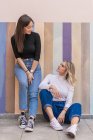 Улыбающиеся позитивные элегантные женщины сидят, опираясь на раздетую красочную стену рядом с тротуаром на улице и смотрят друг на друга — стоковое фото
