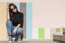 Позитивна розкішна безтурботна жінка в стильному вбранні і сонцезахисних окулярах, спираючись на пофарбовану міську стіну, сидячи на самоті в сонячний день — стокове фото