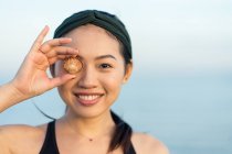 Ásia mulher no esporte camisa olhar para câmara e cobertura olho com mar shell enquanto pé no praia — Fotografia de Stock